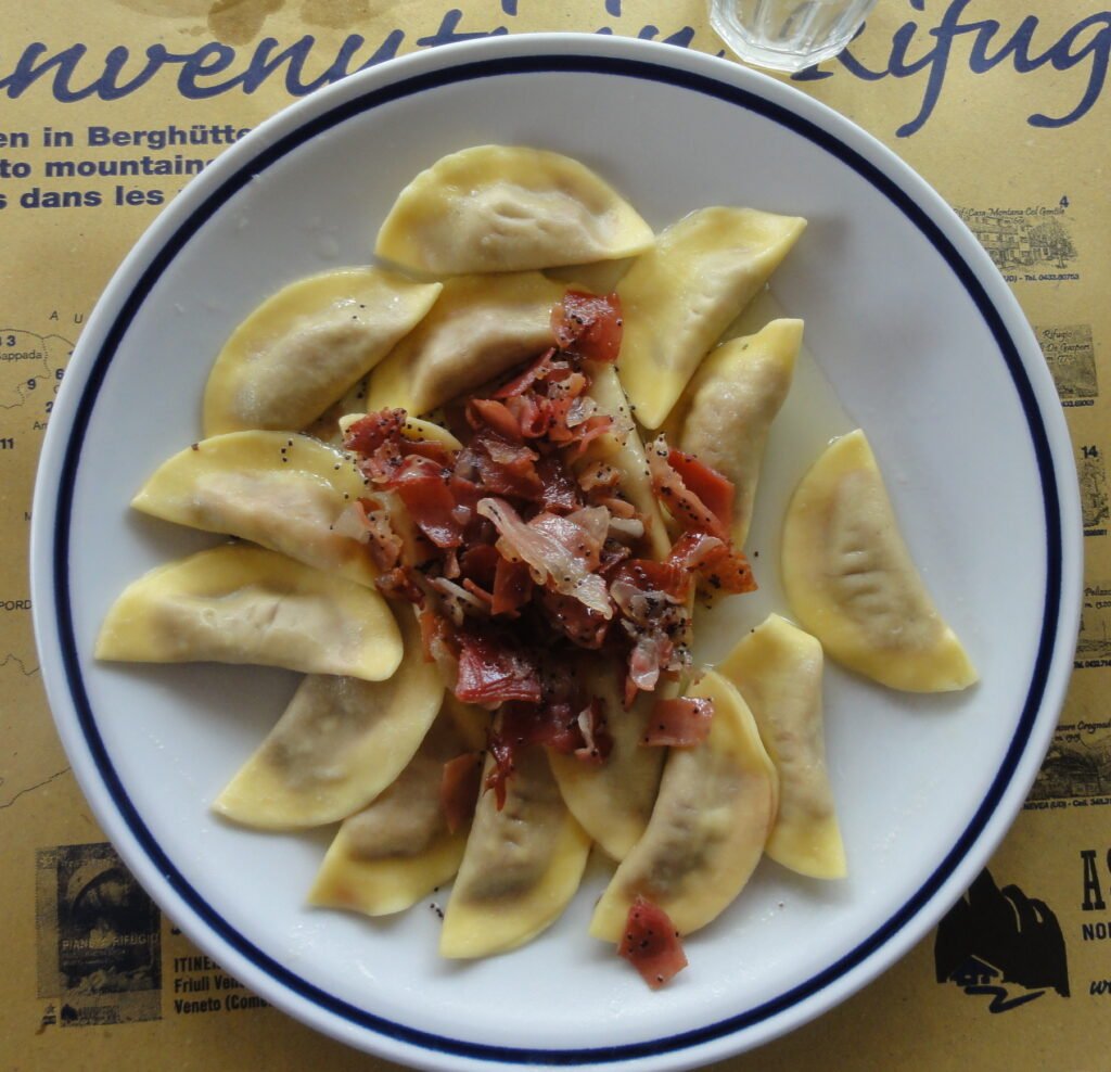 A plate of half-moon casunziei pasta filled with beetroot, topped with crispy speck, butter and poppy seeds