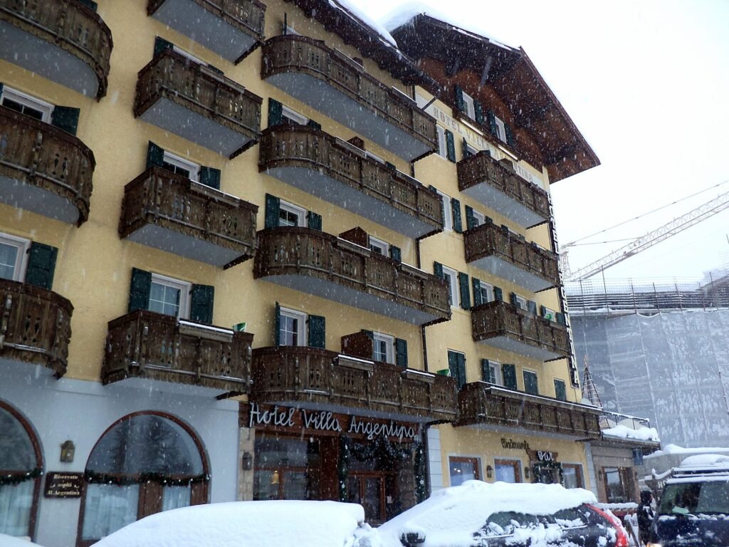 A traditional multi-storey Alpine hotel with wooden balconies and snow-covered cars in front, in Pocol above Cortina