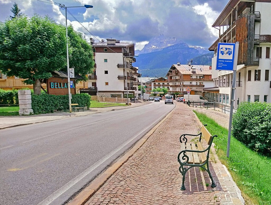 A town bus stop sign beside a bench on a quiet street in Cortina d'Ampezzo, with hotels and mountains behind